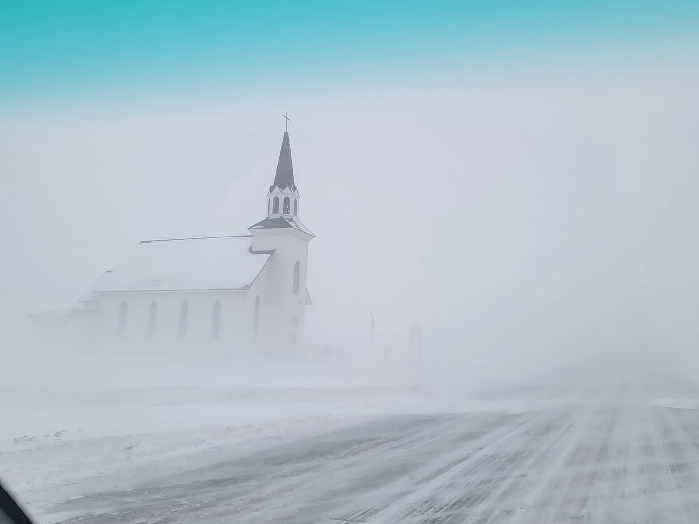 White country Christian church near a road. Snow is drifting so much you can barely see the church, and cannot see far down the road.