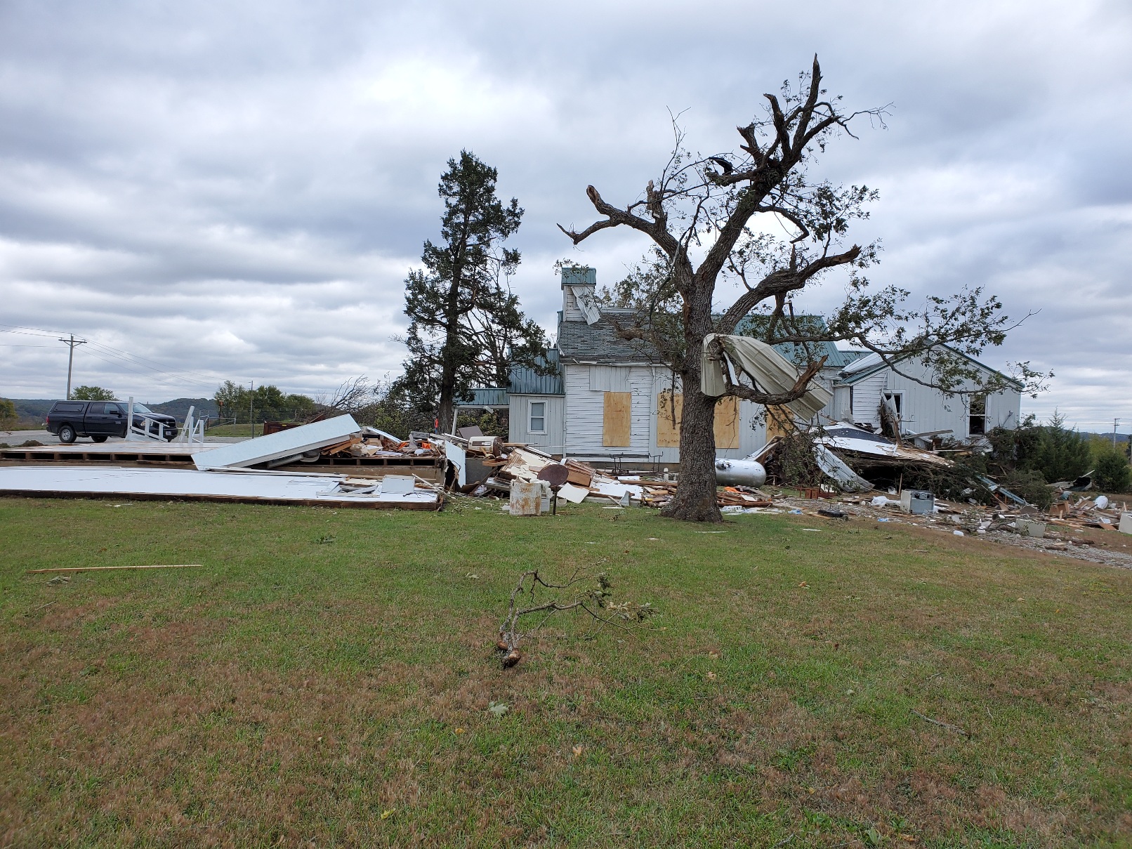 A damaged church stands alone with plywood over the larger side windows. Metal siding is bent and twisted, lying across the ground and stuck in the cedar trees that stand to the side of the church. A one room school house that stood next to the church has been completed destroyed, it's walls pushed over and the contents of the building scattered across the property.