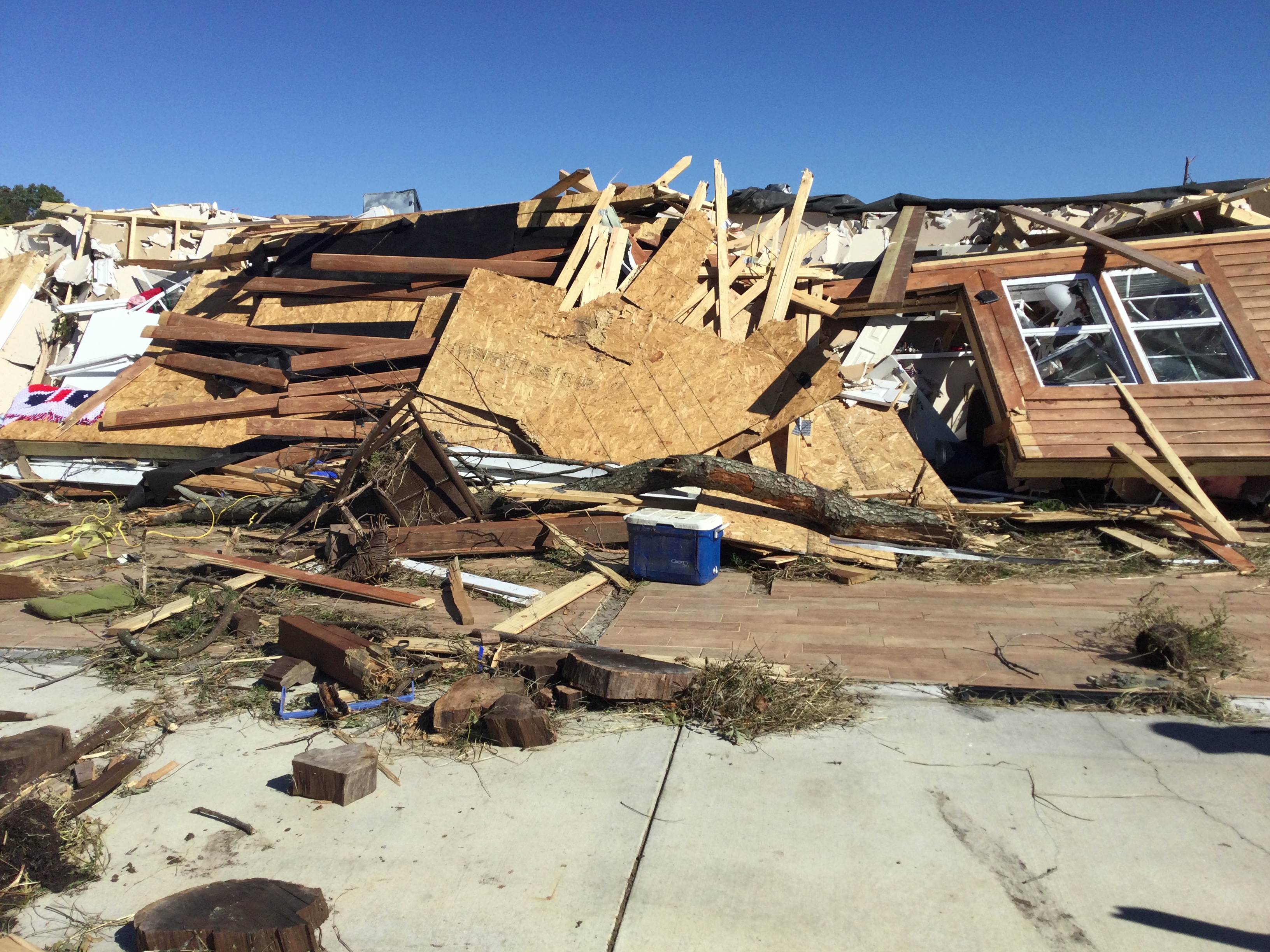 A pile of wood and other building materials lies on top of a wooden floor that used to be where a house stood. A section of wall with the window still in it leans on top of the pile, like the house was blown over and the wall collapsed on top of it. Pieces of splintered wood and other debris litter the foreground.