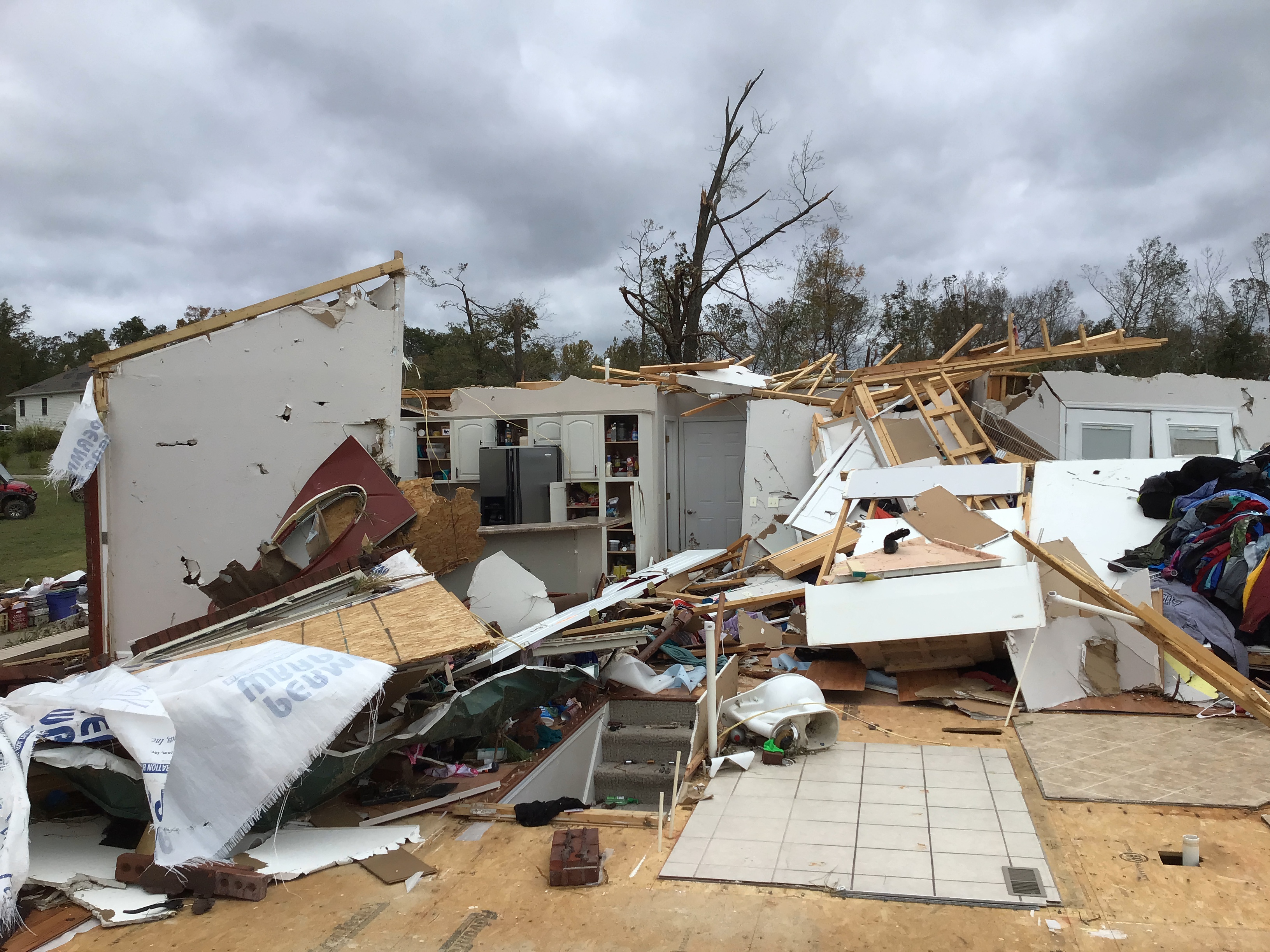 A picture of the inside of a damaged house, standing facing what remains of the kitchen and surrounding areas. The steps to the basement are visible amid the debris of walls and ceilings. The house is completely open to the outside, having lost most of it's exterior walls. This is set against a cloud sky, framed on the bottom by hardwood trees missing their leaves and major branches.