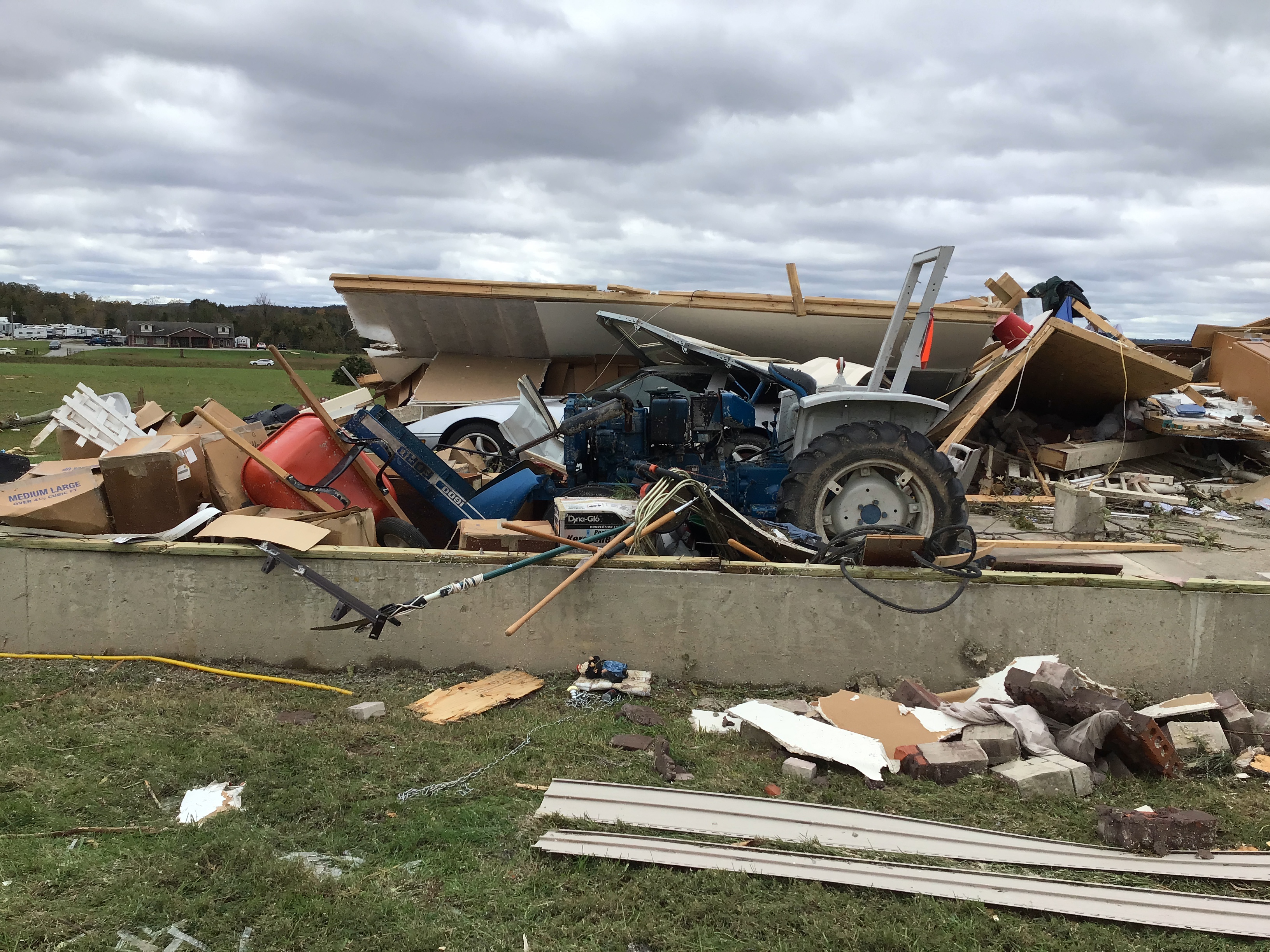 A concrete foundation runs across the bottom third of the image, containing the remains of what appears to be a garage. Portions of a blue tractor, red wheelbarrow, and silver car are just visible among the debris from the walls and ceiling, which appear to have been pushed away from the viewer onto the other side of the building footprint.