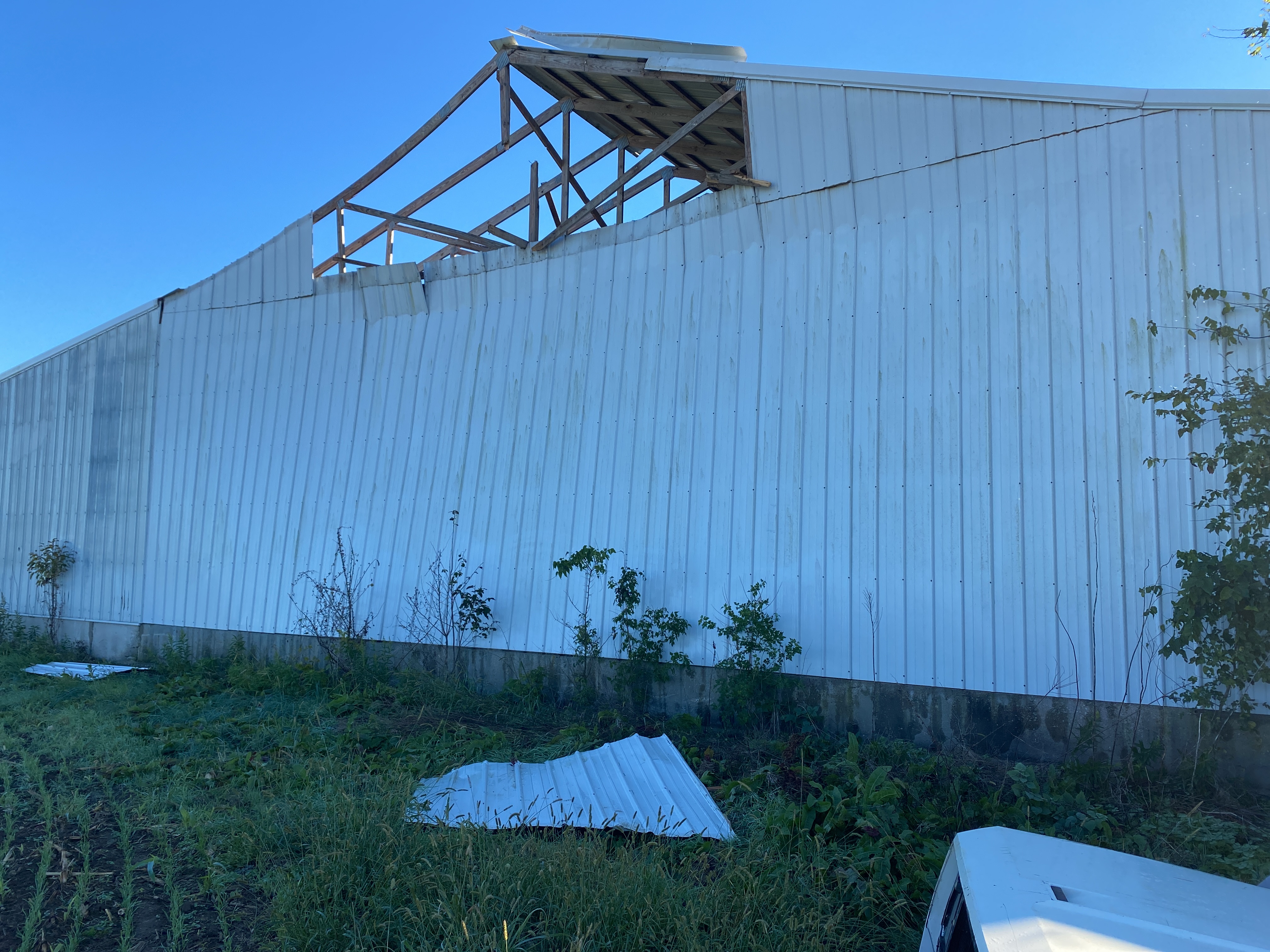 A large outbuilding with white metal siding has lost a portion of it's roof and some of the metal siding, which lies on the grass in the foreground.