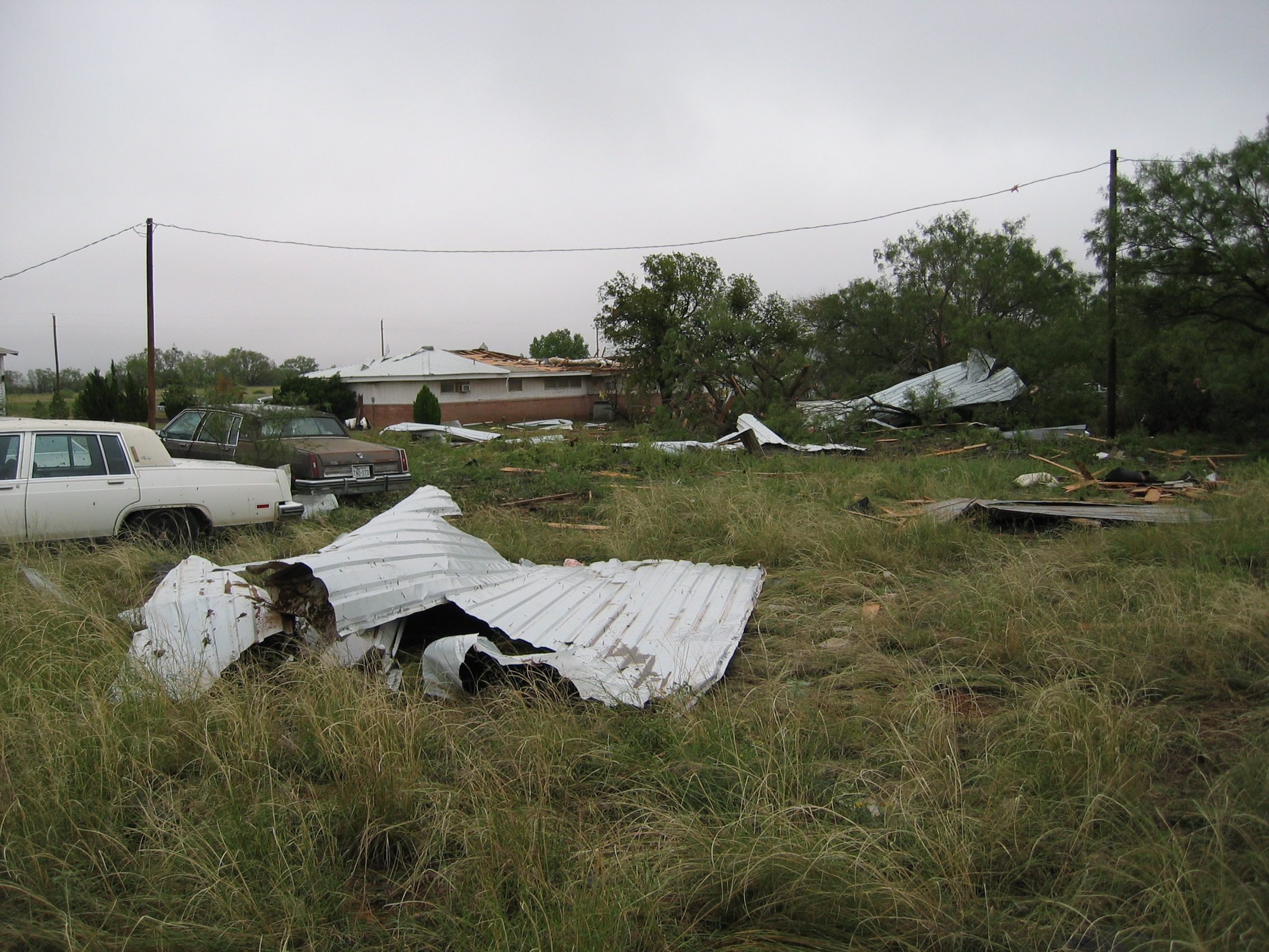 Photo showing more roofing material blown far from the house and a white car rotated 90 degrees from it's parked position