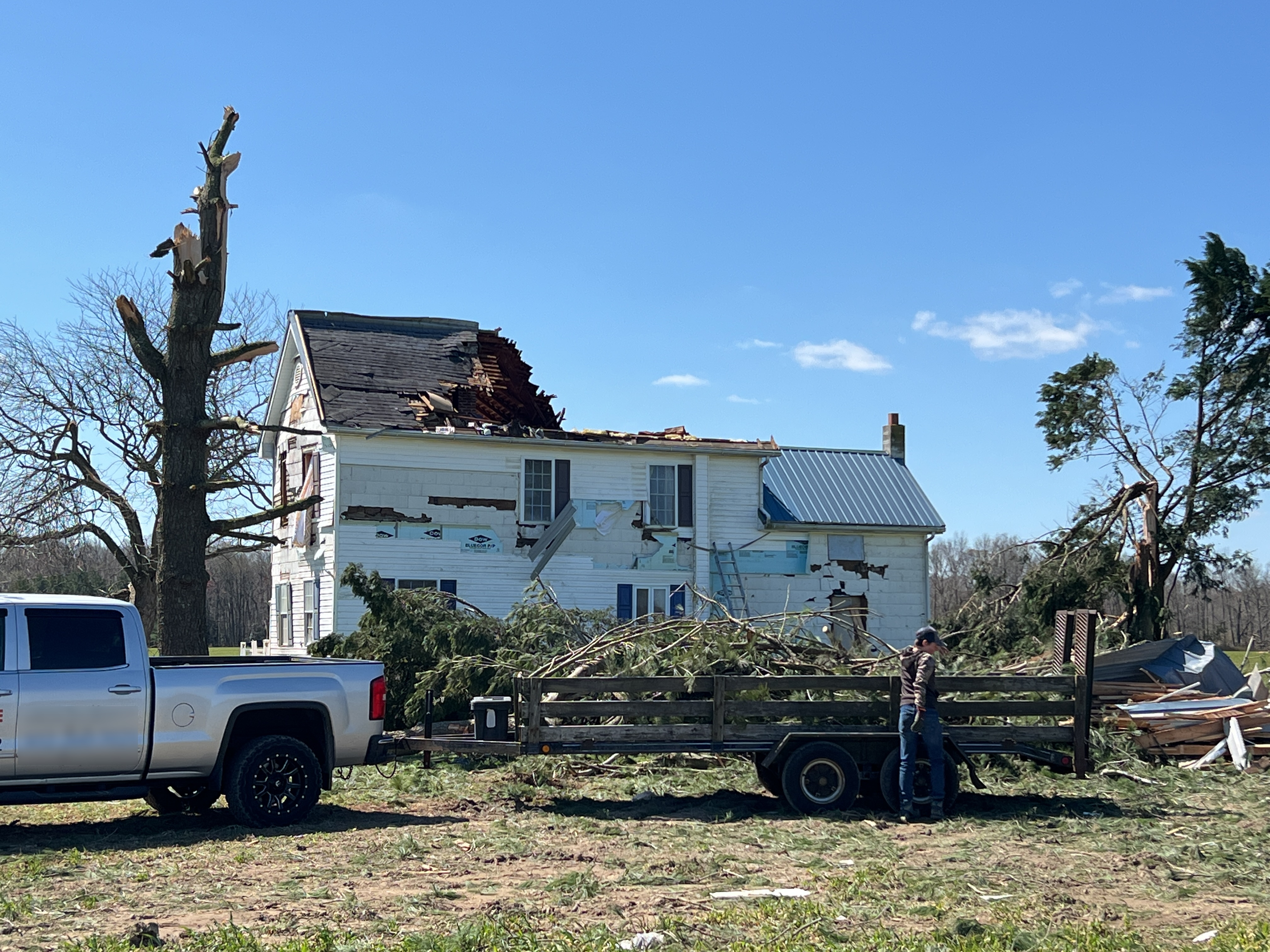 A single family home with large sections of the roof destroyed and much of the siding ripped off. This tornado damaged building is surrounded by snapped tree trunks.