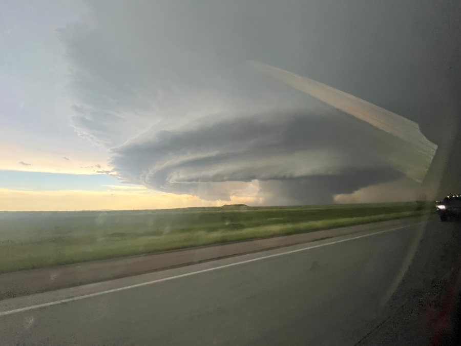 A massive, layered supercell thunderstorm with a rotating updraft dominates the sky over a flat, green landscape. The storm's dark, ominous base and layered cloud structure are prominent against the brighter sky near the horizon. A road is visible in the foreground, with grass on either side, and the view is from inside a vehicle.