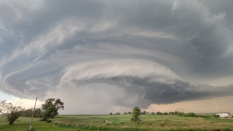 A dramatic photograph shows a large, ominous supercell thunderstorm over a flat, grassy landscape. The cloud formation is immense and circular, with a sculpted, dome-like appearance at its center, surrounded by swirling, layered clouds. A rain shaft is visible beneath the storm's core. The sky is dark and turbulent, with a slight yellowish or orange tint on the horizon to the right, suggesting the time is near sunset. The foreground is a green field with scattered trees and some faint white structures in the distance under the storm.