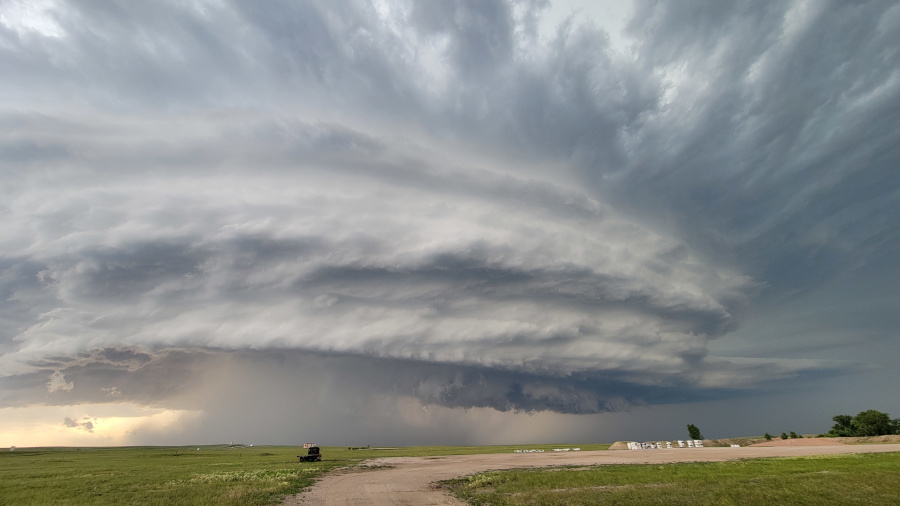 A powerful, multi-layered supercell thunderstorm with a massive, rotating updraft looms over a flat, grassy landscape. Rain and hail shafts are visible under the dark, textured cloud base. In the foreground, there is a dirt road and a small vehicle, with an area of gravel or sand to the right. The sky is filled with dramatic, turbulent clouds, and the scene evokes a sense of both awe and danger.
