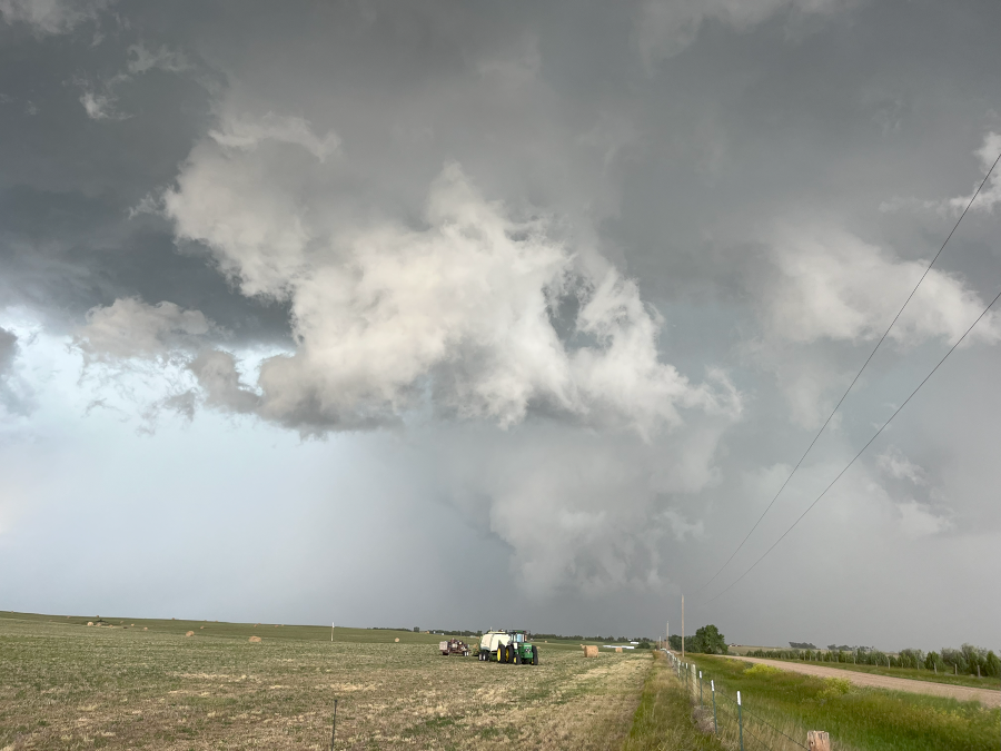 A powerful storm with a rotating updraft and turbulent clouds is shown over a rural landscape. A large, white, cloud formation, possibly a wall cloud or developing funnel, hangs low from the main cloud base. In the foreground, a tractor and another piece of farm equipment are visible in a field with hay bales. The scene is overcast with a sense of impending severe weather.