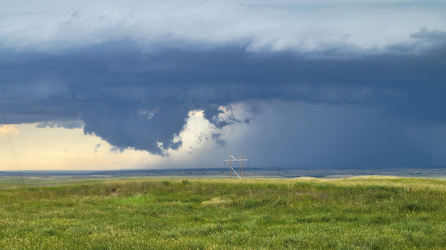 A vast, dark storm cloud dominates the right side of the sky, with a heavy downpour of rain visible underneath. A break in the clouds reveals a brighter patch of sky and some lighter, more turbulent clouds within the storm structure. A lone wooden power line structure stands in the middle of a gently rolling, green field. The horizon is a mix of green hills and a distant, hazy line. The scene is a mix of serene landscape and dramatic weather.