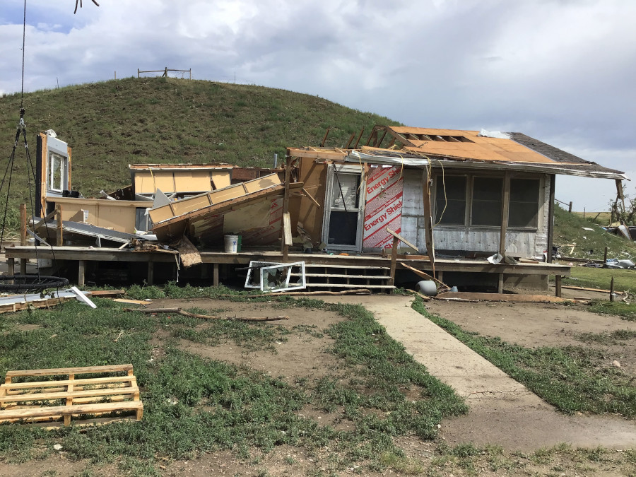  A severely damaged single-story house is shown with its roof and walls partially torn off, exposing the interior. Debris is scattered around the property, and a grassy hill is in the background.