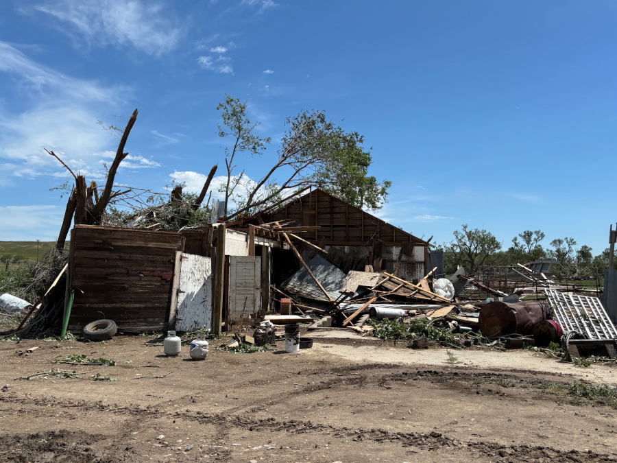 A destroyed wooden barn or outbuilding is surrounded by a large pile of debris, including broken wood, metal siding, and tree limbs. The roof is partially missing and the walls are collapsed.