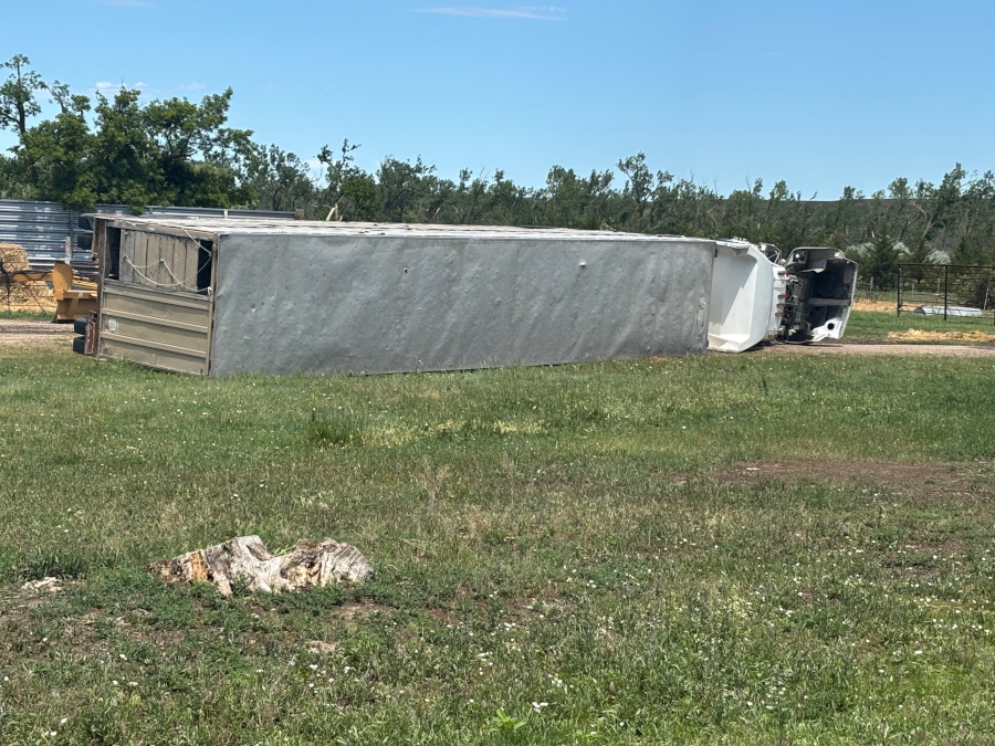 An overturned trailer lies on its side in a grassy field with trees in the background.