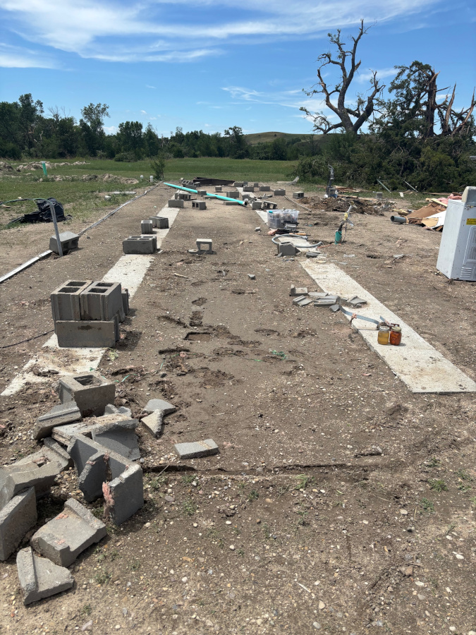  A dirt lot is covered with scattered cinder blocks, pieces of concrete, and other debris. In the background, a large, broken tree is visible under a blue sky.